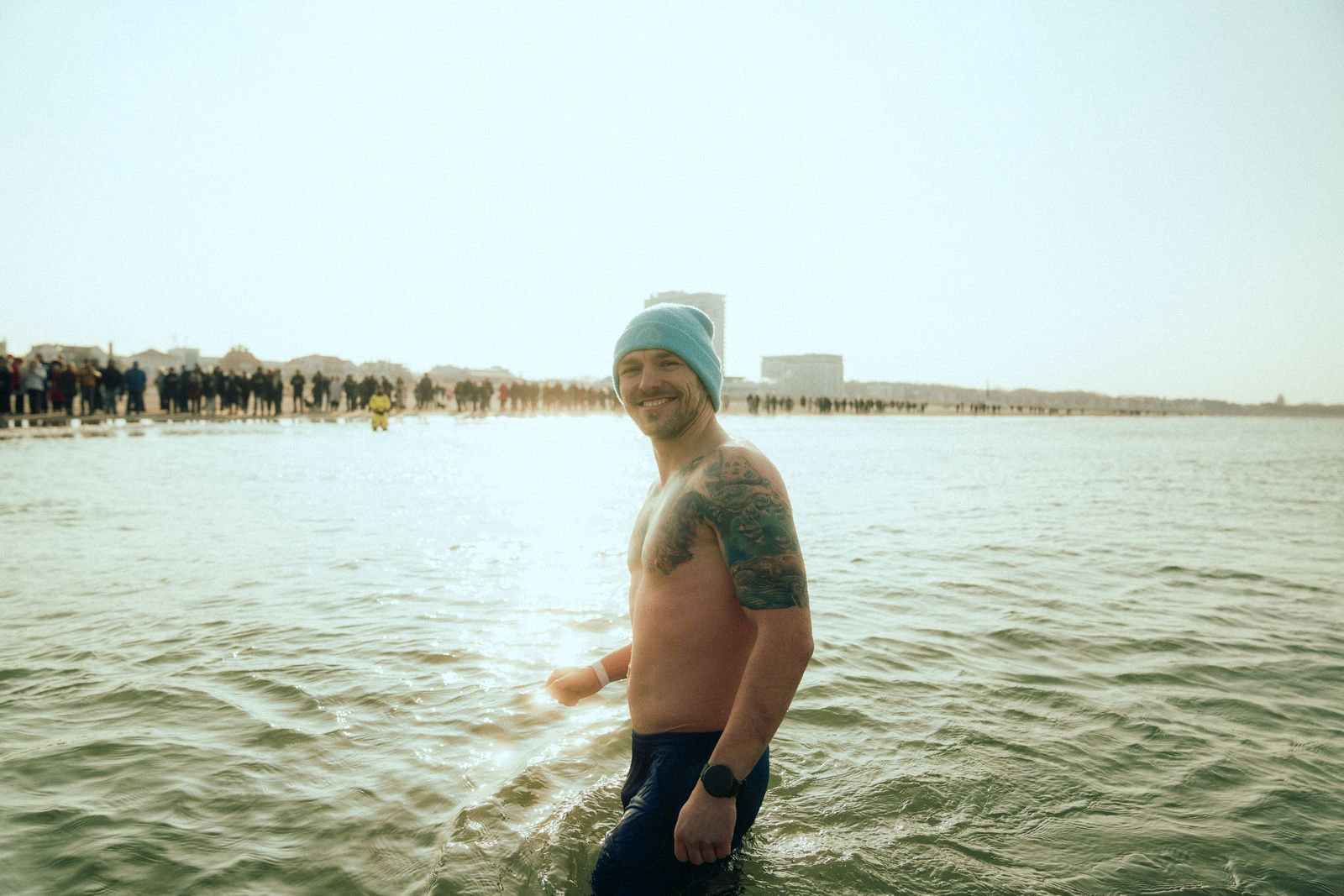 Phillipp Risch steht lächelnd im kalten Meerwasser, im Hintergrund Zuschauer am Strand